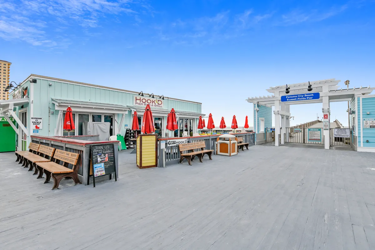 A bustling outdoor restaurant with vibrant red umbrellas and benches, adjacent to the entrance of a scenic pier. The inviting space suggests opportunities for gourmet dining and leisurely strolls by the waterfront.