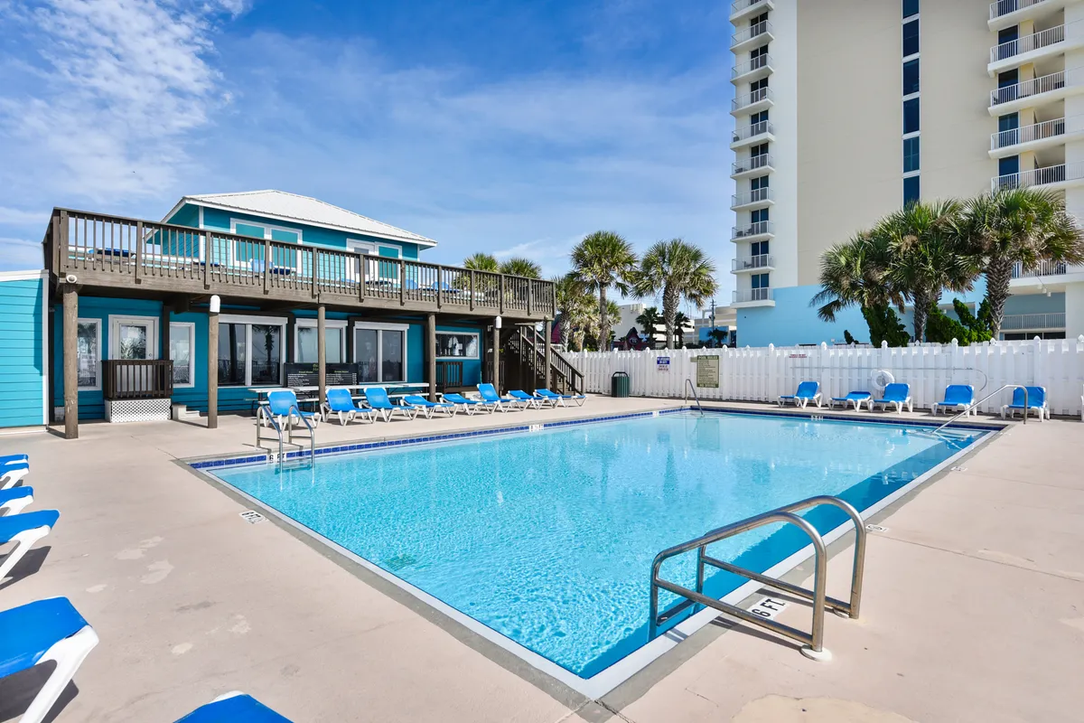 A sunlit swimming pool and deck area framed by palm trees, with bright blue skies overhead. Lounge chairs and outdoor spaces hint at opportunities for sunbathing, swimming, and casual group gatherings. There is a sense of vibrancy and relaxation, ideal for families enjoying leisurely afternoons together.