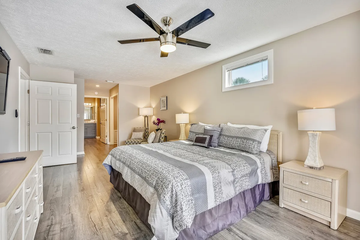 A spacious master bedroom featuring a king-size bed with patterned bedding, an armchair and side table, wall-mounted flat-screen TV, decorative lamps, and subtle floral accents. The room exudes a serene, comfortable atmosphere with ample space to relax or prepare for the day.
