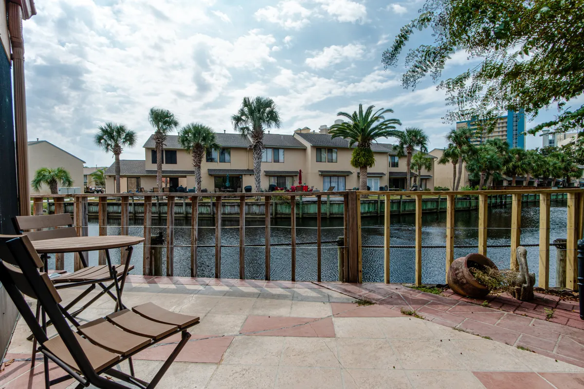 A serene lagoon-front patio features a small outdoor table and chairs set on a wooden balcony, overlooking calm lagoon waters that reflect the sky and nearby resort buildings. A decorative clay pot with lush plants adds a touch of greenery, creating a tranquil space for morning coffee or evening relaxation.
