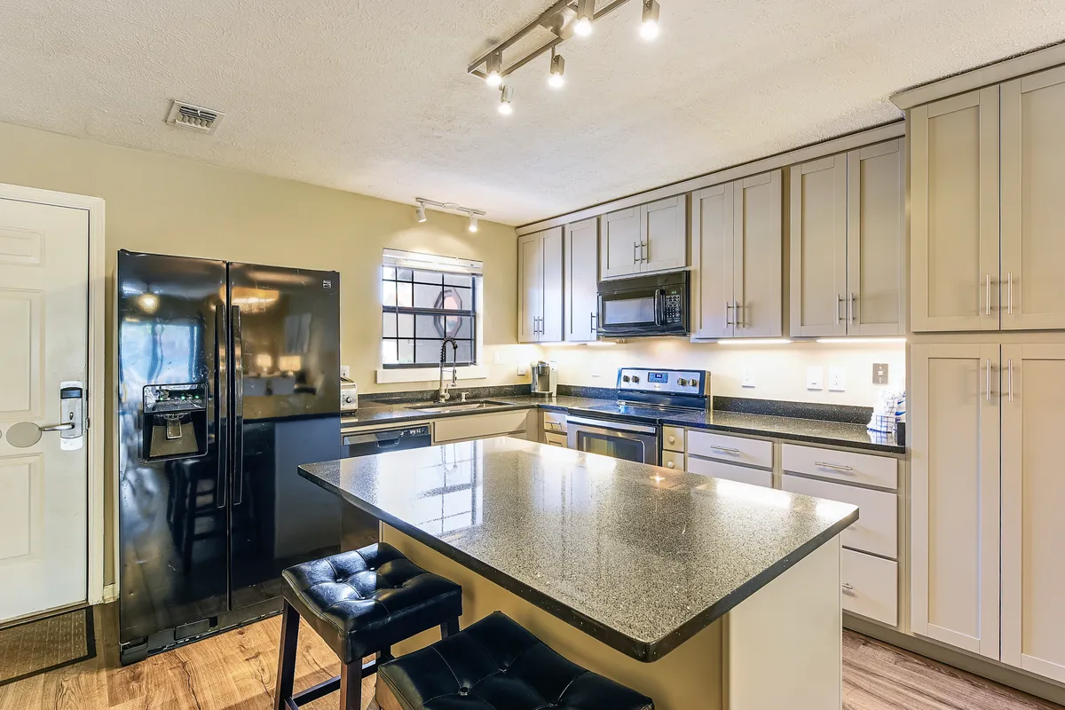 A spacious kitchen with a granite-topped island and black cushioned stools for casual seating. The black refrigerator features a water dispenser, and the room is equipped with ample light gray cabinetry and modern hardware, creating a welcoming and practical setting for cooking and socializing.