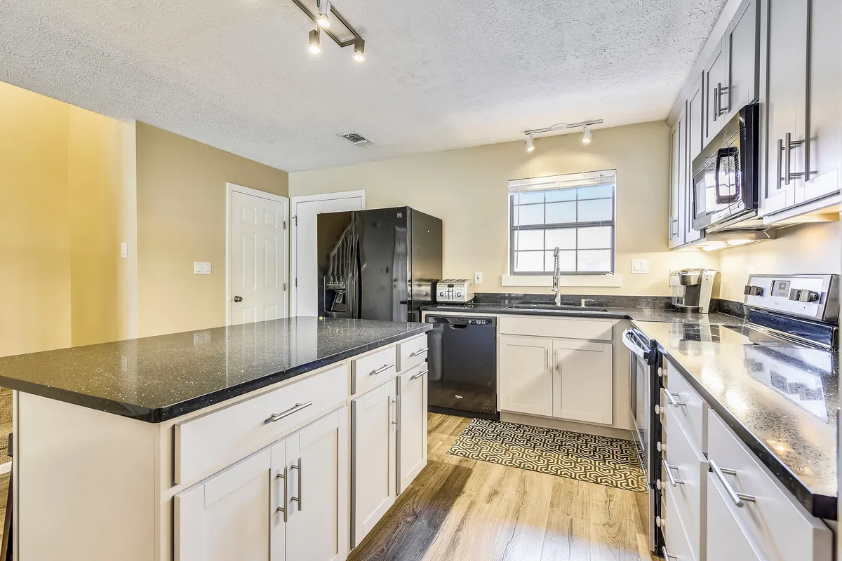 A bright and inviting kitchen featuring a large kitchen island with a sleek black countertop, a black refrigerator and dishwasher, and a window above the sink that fills the space with natural light. The practical layout allows for both meal prep and casual dining or conversation around the island.
