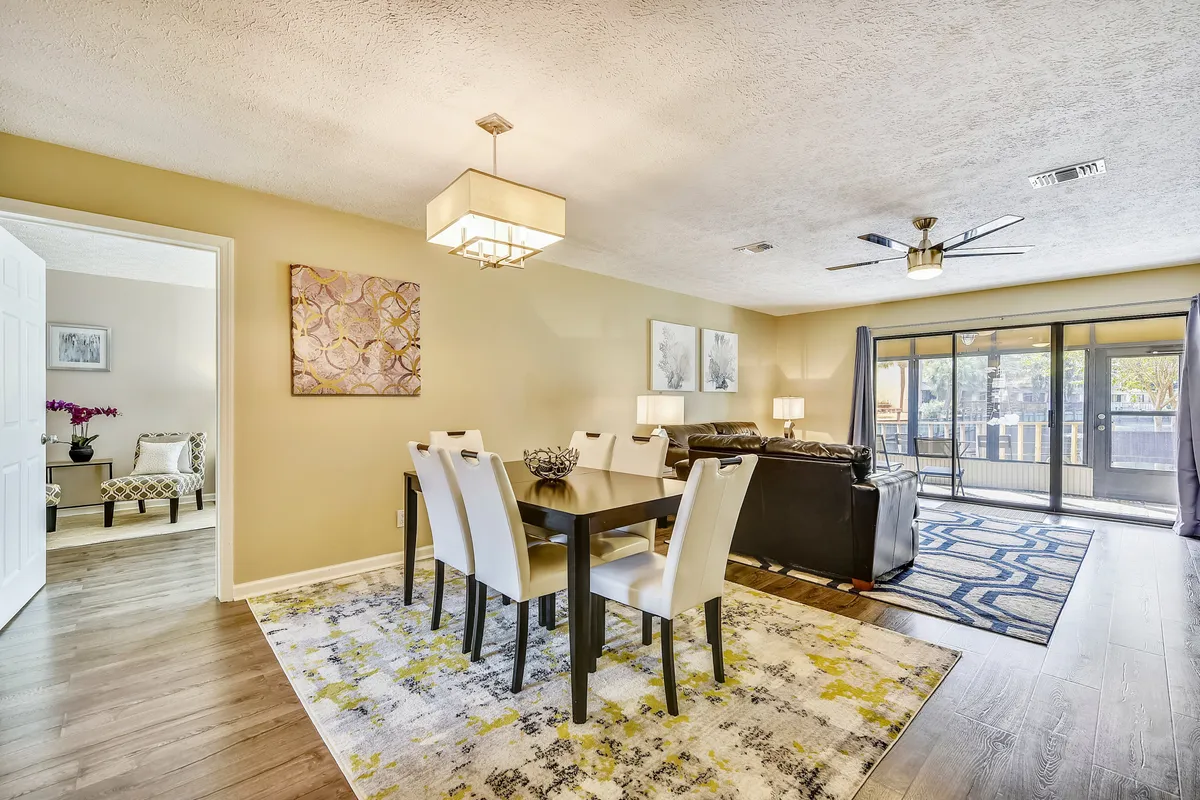 This image captures an inviting open-concept living and dining space. The dining table with six chairs is positioned for communal meals, while the adjacent living area features leather sofas, decorative rugs, and a large sliding glass door leading to a patio. The space is bright and welcoming, ideal for both relaxation and group activities.