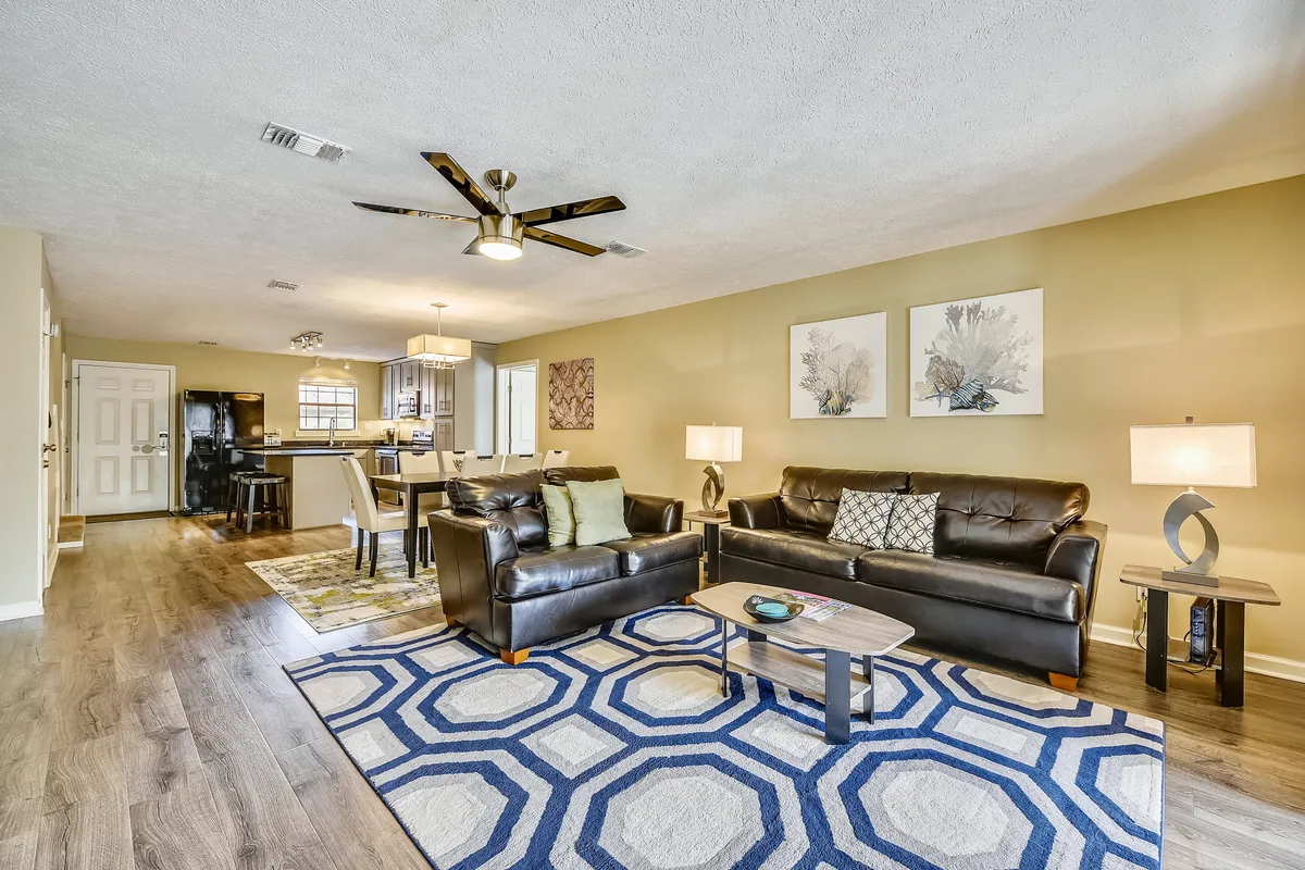 A comfortable living room featuring black leather sofas with decorative cushions, a geometric patterned blue and white area rug, a wooden coffee table, and an open view into the kitchen and dining area in the background. This layout supports both lounging and easy socialization across the main communal spaces.