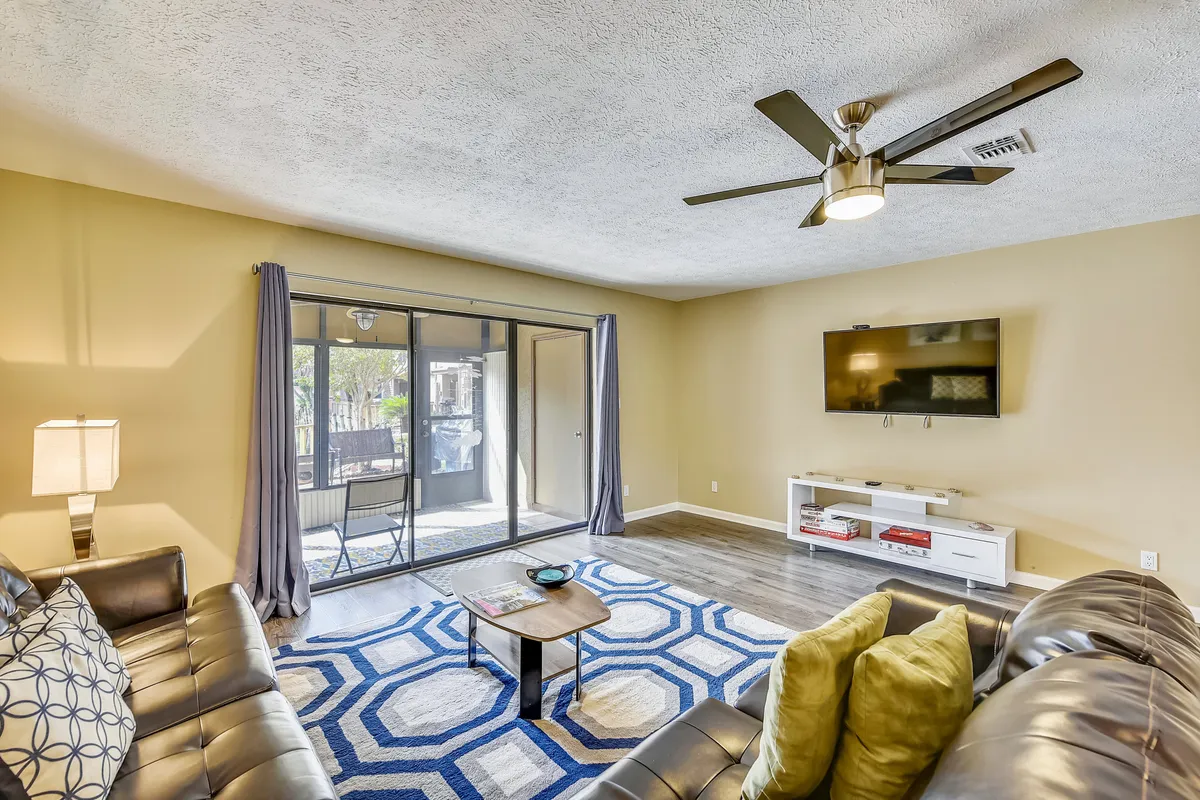 The living area is equipped with leather sofas, a wall-mounted flat screen television, and a large sliding glass door that leads to a screened-in patio. Decorative pillows and a geometric area rug add style and comfort, creating a welcoming environment for entertainment and relaxation.