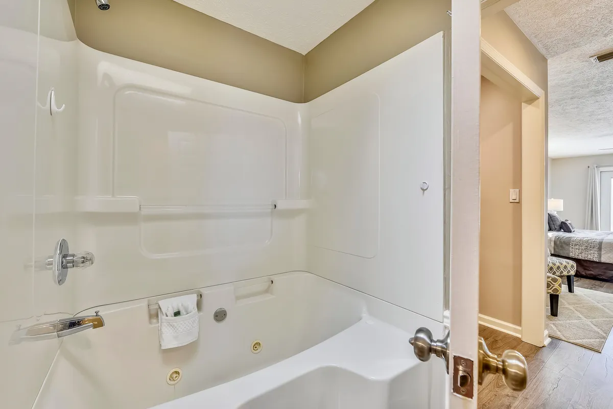 A bright and calming bathroom with a white bathtub equipped with built-in jets, a chrome faucet and showerhead, and an open door leading to the bedroom. The space is designed for relaxation after an active day.
