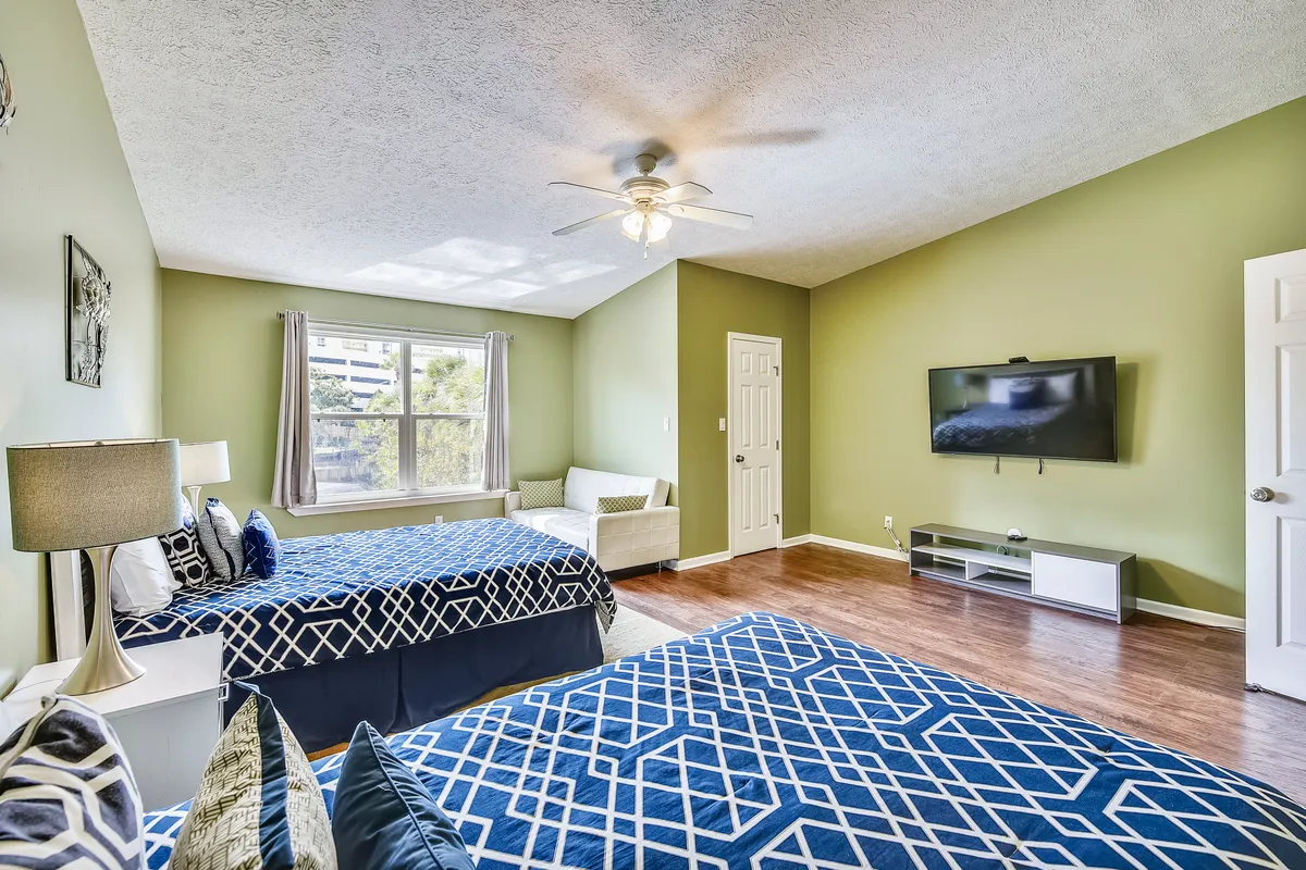 A restful bedroom scene with two beds in blue geometric patterned bedding, a flat-screen television mounted on a green accent wall, a white loveseat near the window, and a ceiling fan with a light fixture. The room is airy, modern, and well-appointed for downtime.