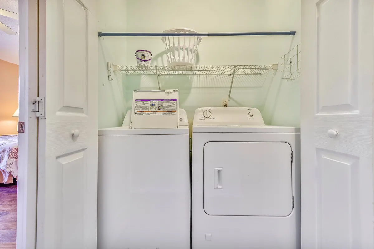 A dedicated laundry nook with a white washing machine and dryer, topped by a wire shelf holding a laundry basket and detergent. The area is clean, organized, and accessible, illustrating the ease of keeping up with laundry needs during a stay.
