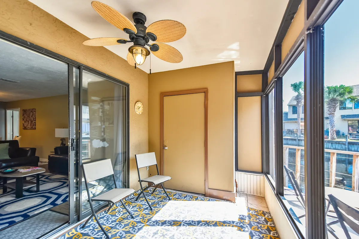 A bright, screened-in porch area features a colorful patterned tile floor, a ceiling fan with leaf-shaped blades, and expansive windows framing the tranquil canal and neighboring houses. Comfortable seating invites you to relax and take in the waterfront view, while the screen enclosure ensures a bug-free, breezy experience.