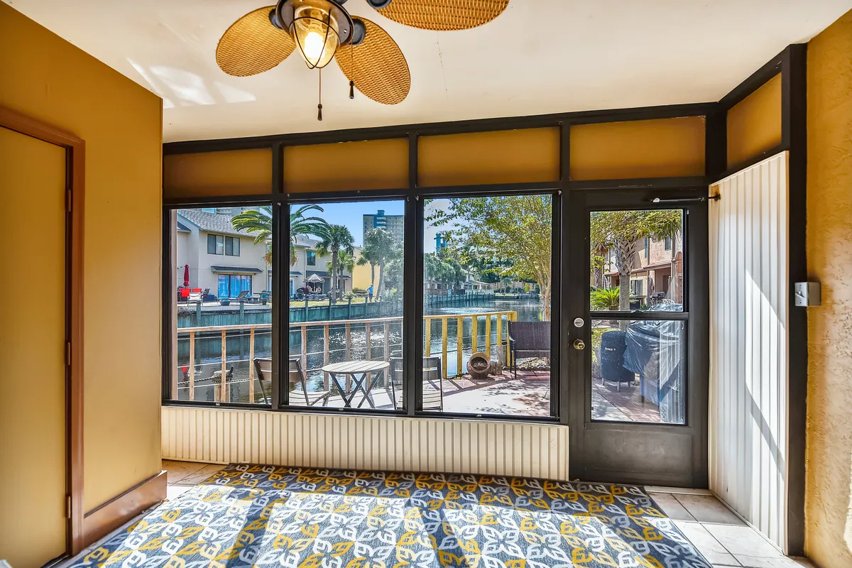 A cozy interior space opens onto large screened windows overlooking the canal, with a ceiling fan featuring woven palm leaf blades and a vibrant tile floor. Through the windows, a small outdoor seating area is visible, reinforcing the seamless blend of indoor comfort and outdoor vistas.