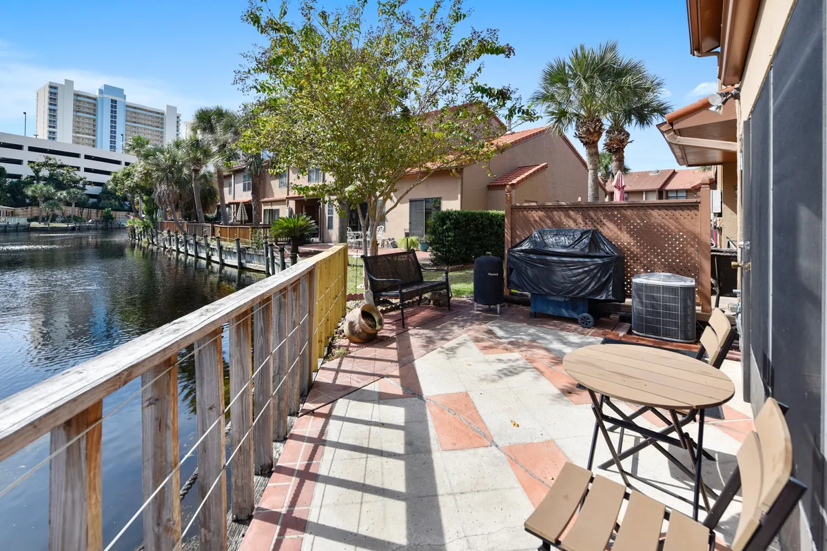A peaceful lagoon-front patio overlooks the canal, featuring an outdoor grill with a black cover, a small round dining table with chairs, and bench seating shaded by a tree. The wooden railing and waterway create an inviting setting for outdoor dining or socializing while enjoying tranquil water views.