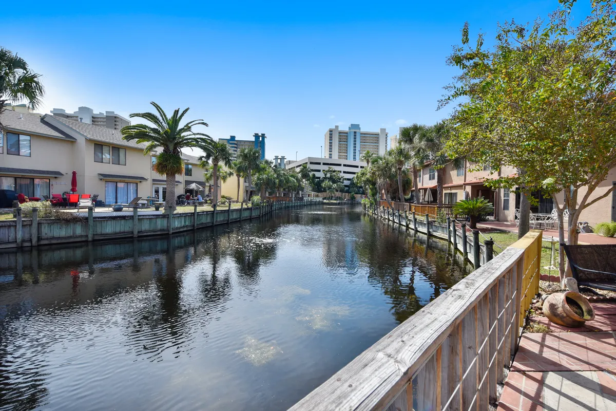 A tranquil canal lined with modern townhouses, each featuring private patios overlooking the calm water. Palm trees and lush greenery frame the scene, and the foreground shows a comfortable patio area with a wooden railing, perfect for group gatherings or quiet conversation while watching boats or wildlife.