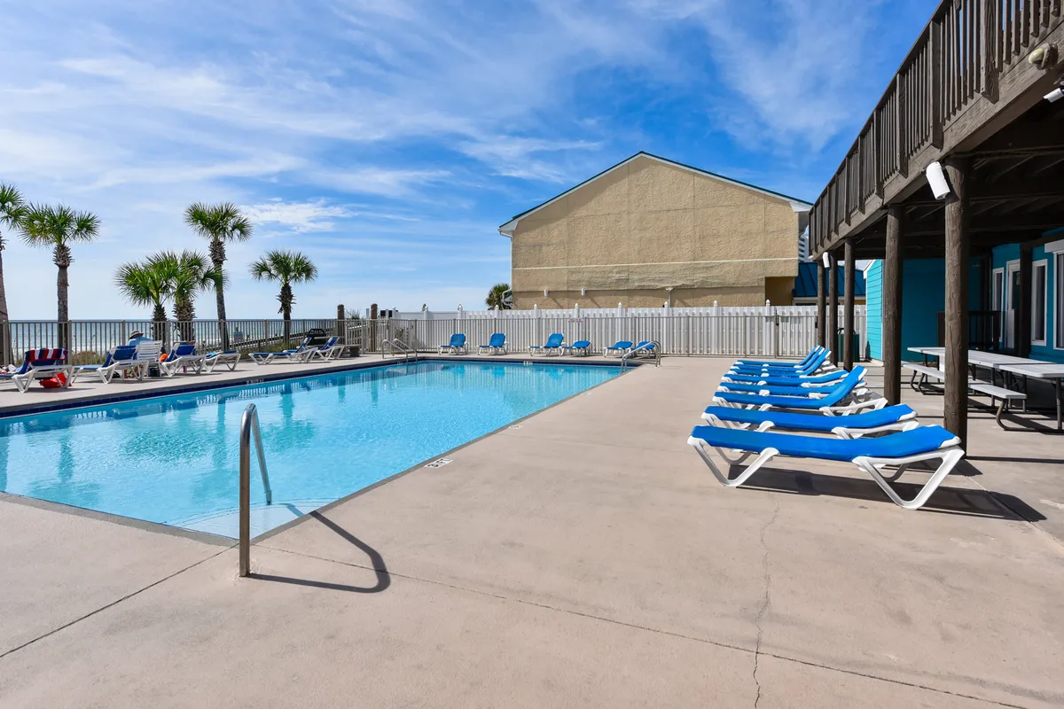 A large rectangular beachfront swimming pool is surrounded by rows of blue lounge chairs and tall palm trees. Beyond the pool fence, the ocean is visible, reinforcing the resort’s proximity to the beach and luxury amenities designed for relaxation and social gatherings.