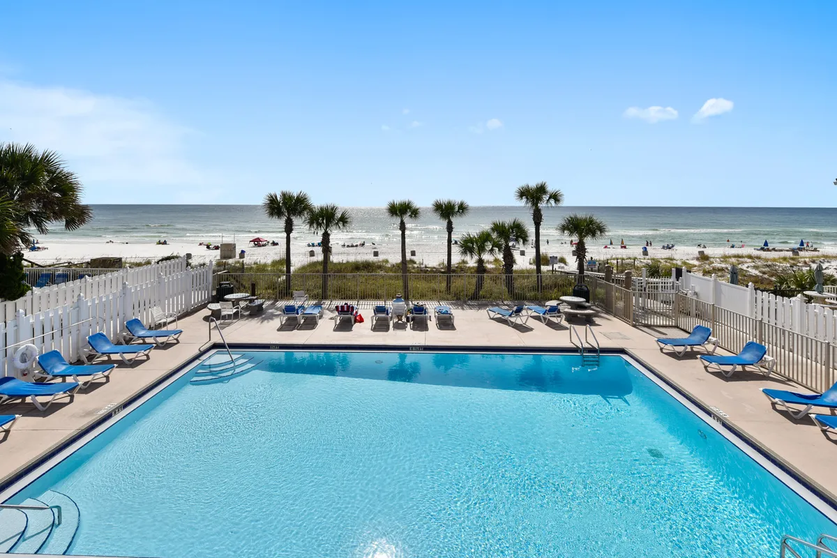 A large rectangular swimming pool with clear blue water sits adjacent to a white picket fence, which separates the pool area from direct beach access. Rows of palm trees line the sandy beach just beyond the fence, and the Gulf of Mexico is visible in the background. Blue lounge chairs are neatly arranged for sunbathing and relaxation.