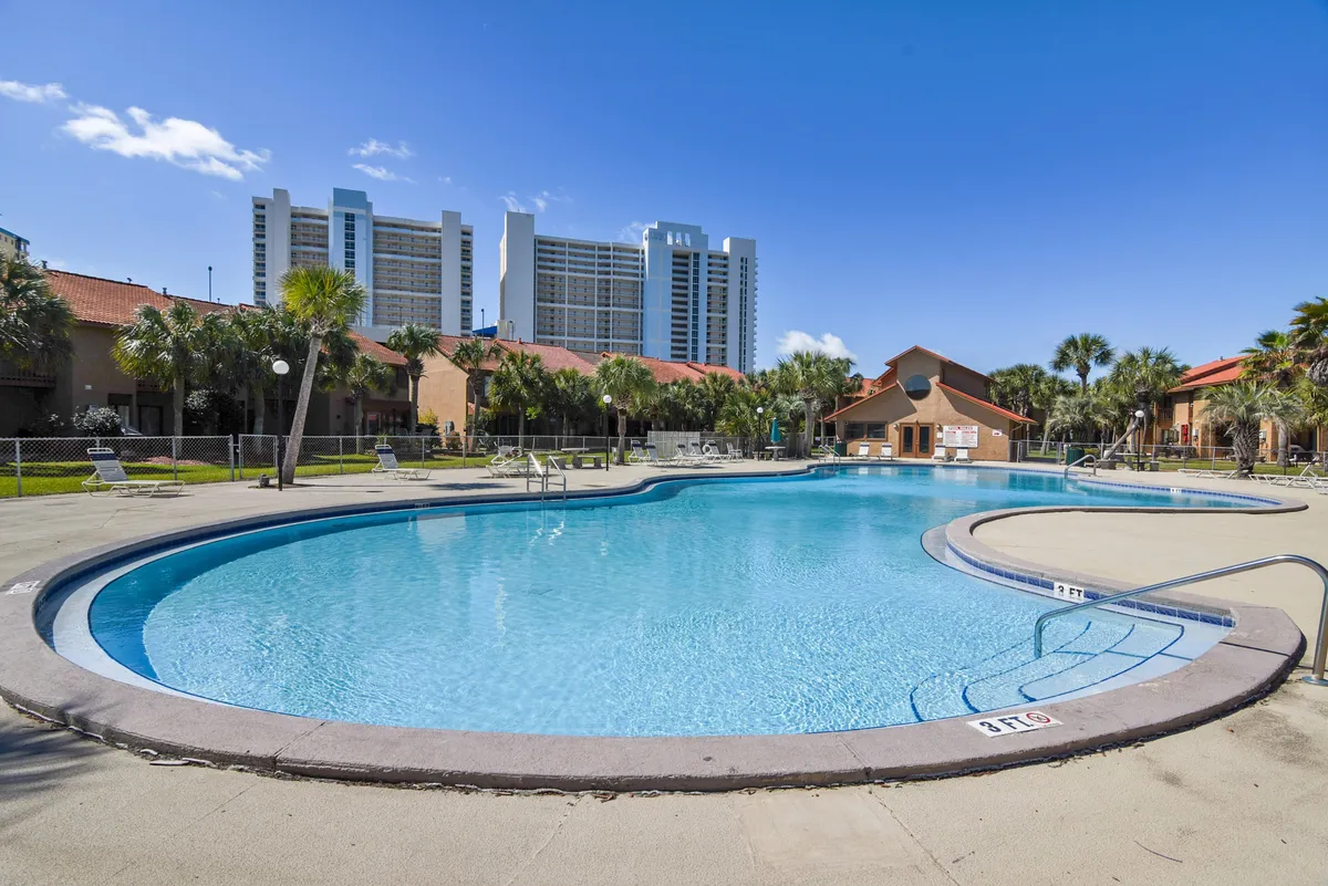 A vibrant outdoor community swimming pool surrounded by lounge chairs and resort buildings, with palm trees adding a tropical vibe. Families and groups can gather for swimming, sunbathing, or socializing poolside, with tall condos in the background hinting at the resort's central location.