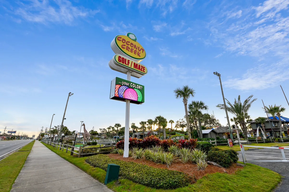 The Coconut Creek Golf/Maze sign anchors a playful outdoor scene featuring whimsical mini-golf course structures and a giraffe statue surrounded by palm trees and tropical landscaping. Families can be imagined enjoying a round of mini-golf in this fun, inviting setting.