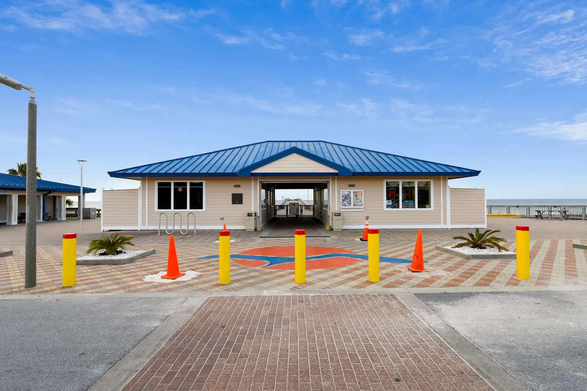 A central pavilion building with a bright blue roof leads to a walkway adorned with colorful painted designs on the brick pavement. Yellow bollards and orange cones add pops of color, while the beach and sparkling ocean are visible in the background. The scene is lively and inviting, suggesting easy access to beach adventures.